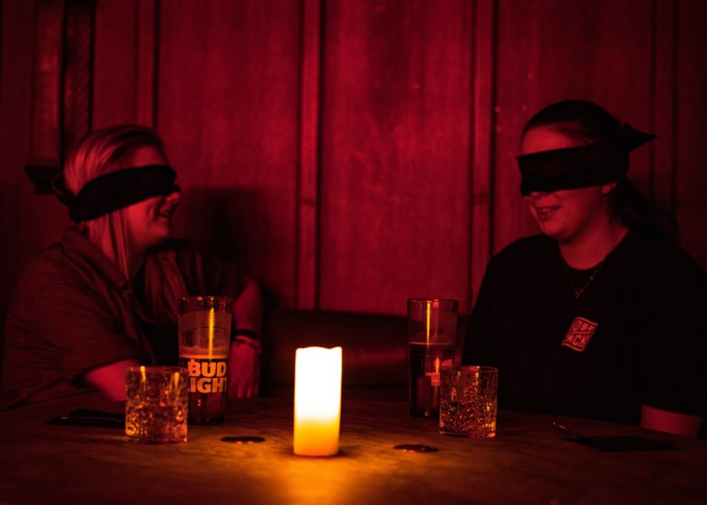 two women sitting at a candlelit table with a blindfold on for the dining in the dark experience