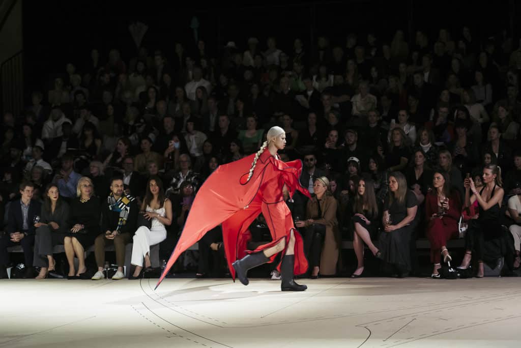 a model in red strutting down the runway in front of a crowd at Melbourne Fashion Week