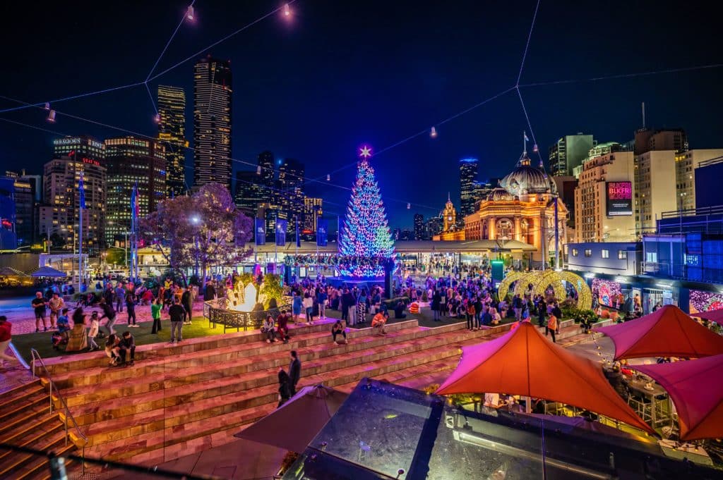 A tall Christmas tree in Fed Square, one of the main locations of the Melbourne Christmas Festival. The tree is lit up at night with Flinders Street Station in the background