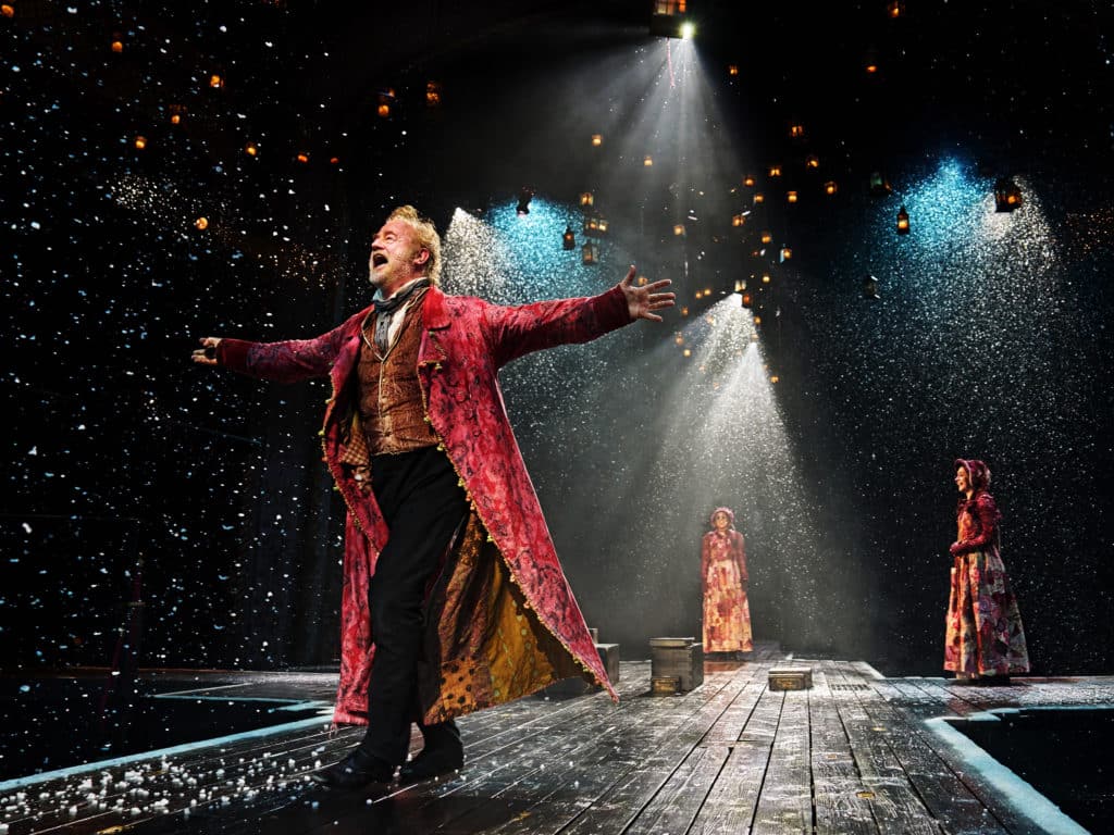 Owen Teale as Scrooge on stage with his arms spread wide open at the 2022 performance of A Christmas Carol at The Old Vic