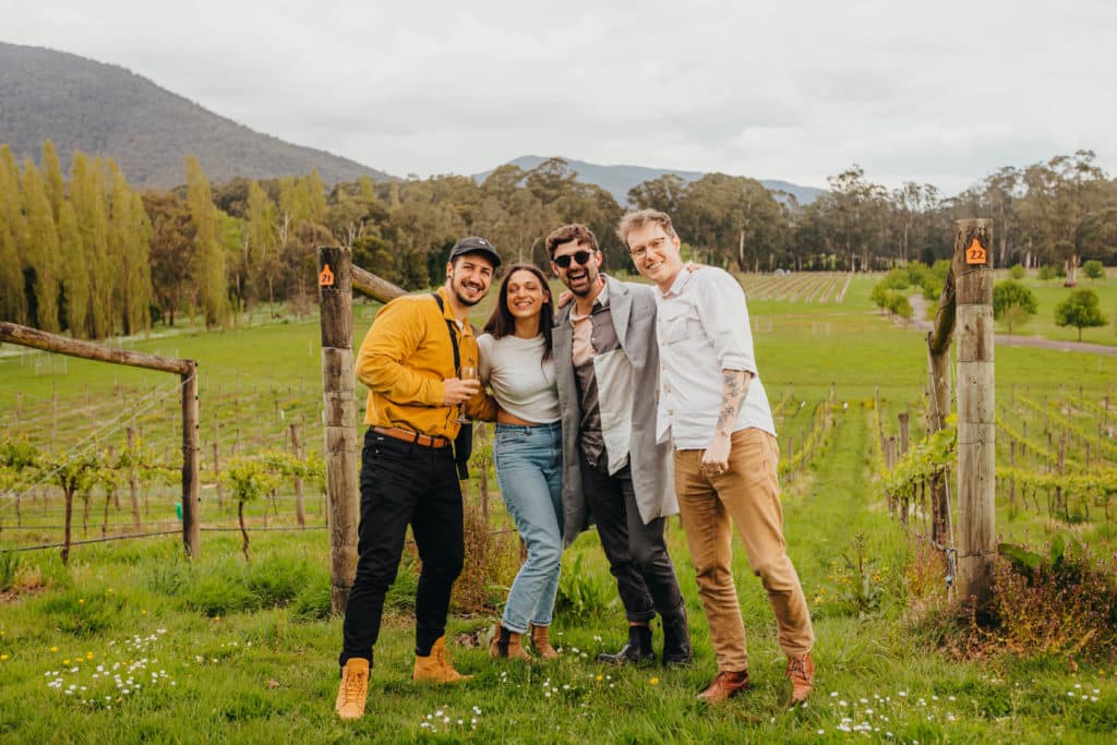 Four people posing with wine glasses in front of a vineyard at Shedfest