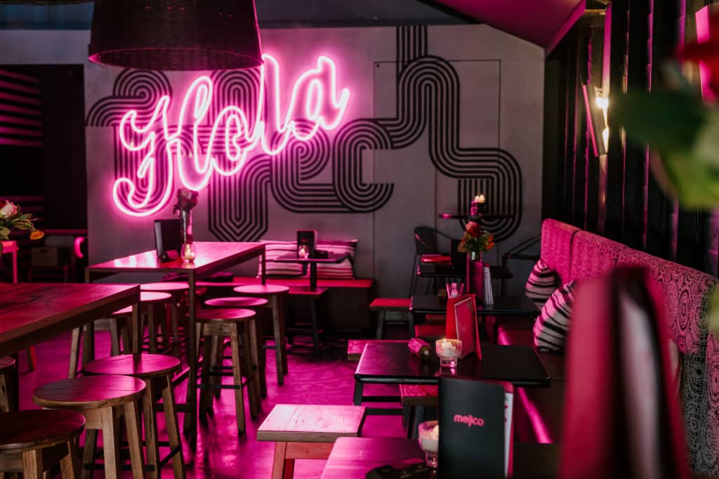 A late-night bar illuminated by a pink neon sign that reads 'Hola'.