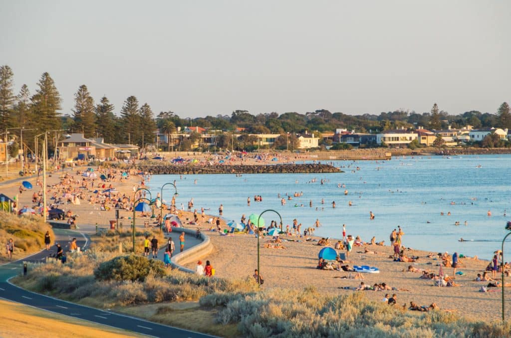 image of elwood beach in melbourne showing people on the sand with beach umbrellas, with winding promenade on the left and water on the right, beach snaking off into the distance