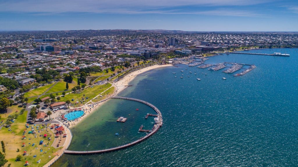 aerial view of eastern beach in geelong showing waterfront including green parks and playgrounds, shark-proof enclosure, kiddy pool and open beach with housing in the background
