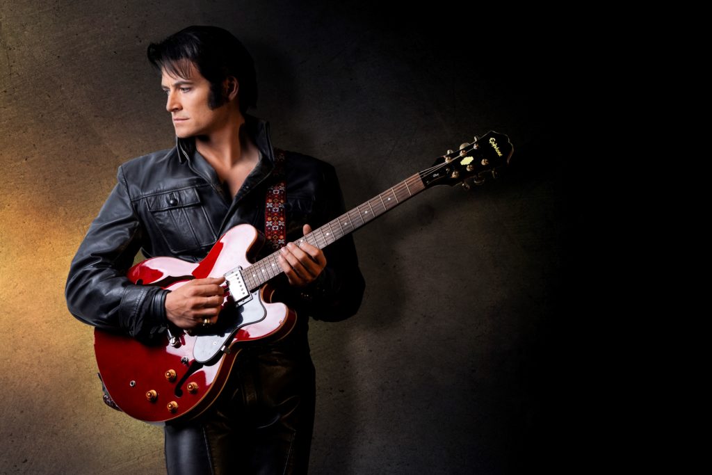 Rob Mallett as Elvis Presley, holding a guitar and looking off to the side while standing in front of a black background