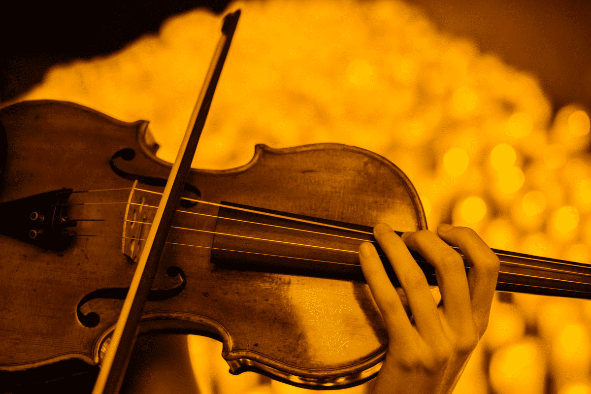 A close-up of a violin with the hand playing it visible and the blurry glow of candlelight in the background.