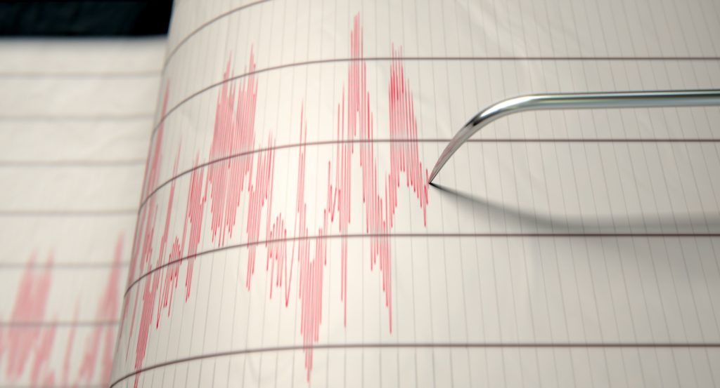 A close-up of a seismograph machine needle drawing a red line on graph paper depicting seismic and earthquake activity