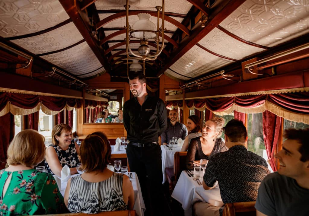 a man talking to seated guests in a dining carriage on the Puffing Billy train