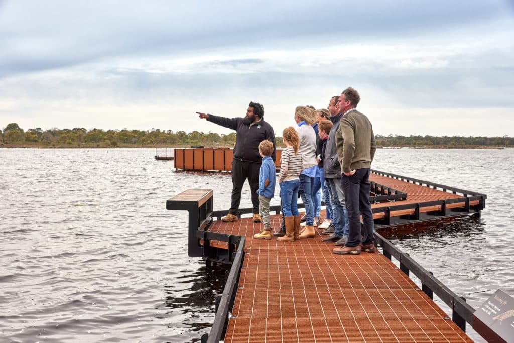 tourists standing on a jetty on a lake, listening while a guide points out something in the distance