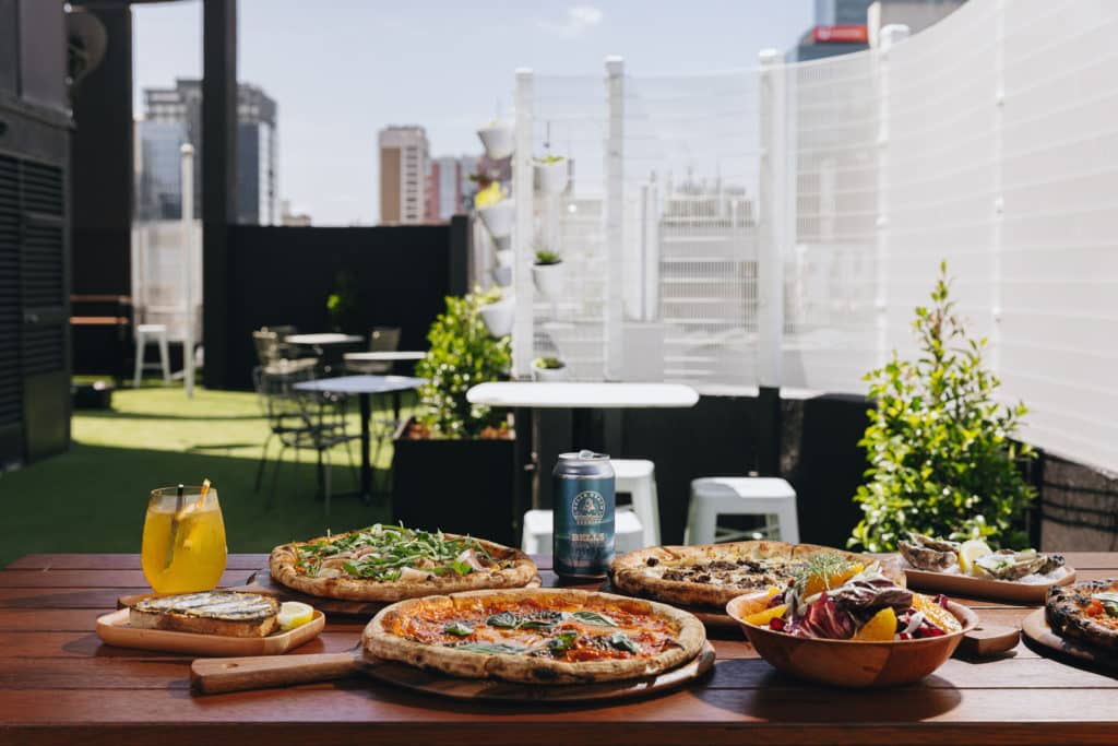 pizza and drinks on a table with a bit of Melbourne skyline in the background at Blossom Rooftop Bar
