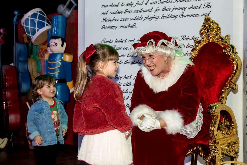Mrs Claus listening and smiling to a little girl while a young boy hovers nearby