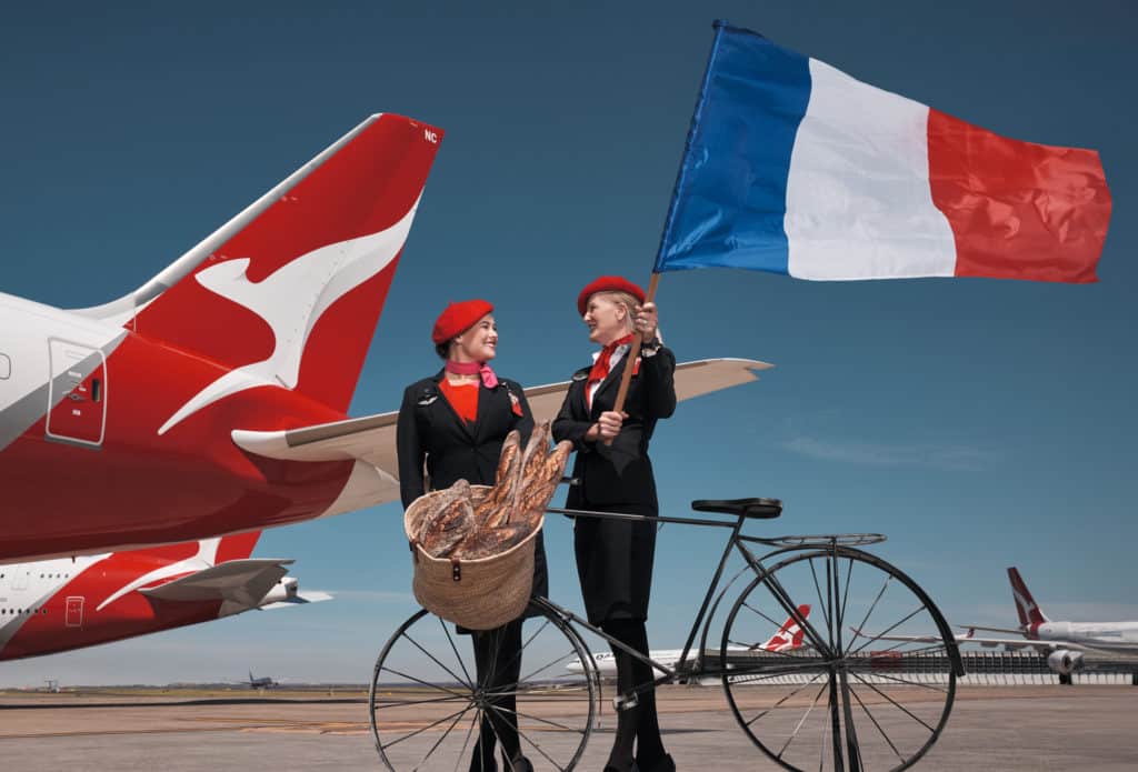 two Qantas flight attendants holding a French flag with a Qantas plane tale behind them. A bicycle and a basket of baguettes is in front of them.