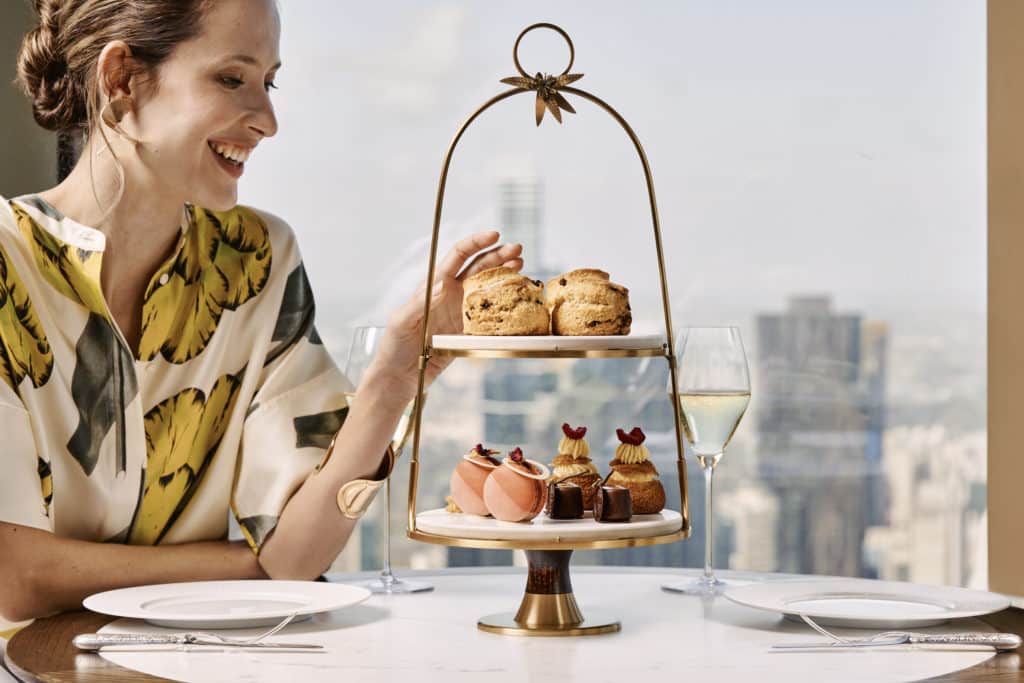 a woman enjoying afternoon tea at The Ritz-Carlton, with a view of Melbourne's skyline behind her